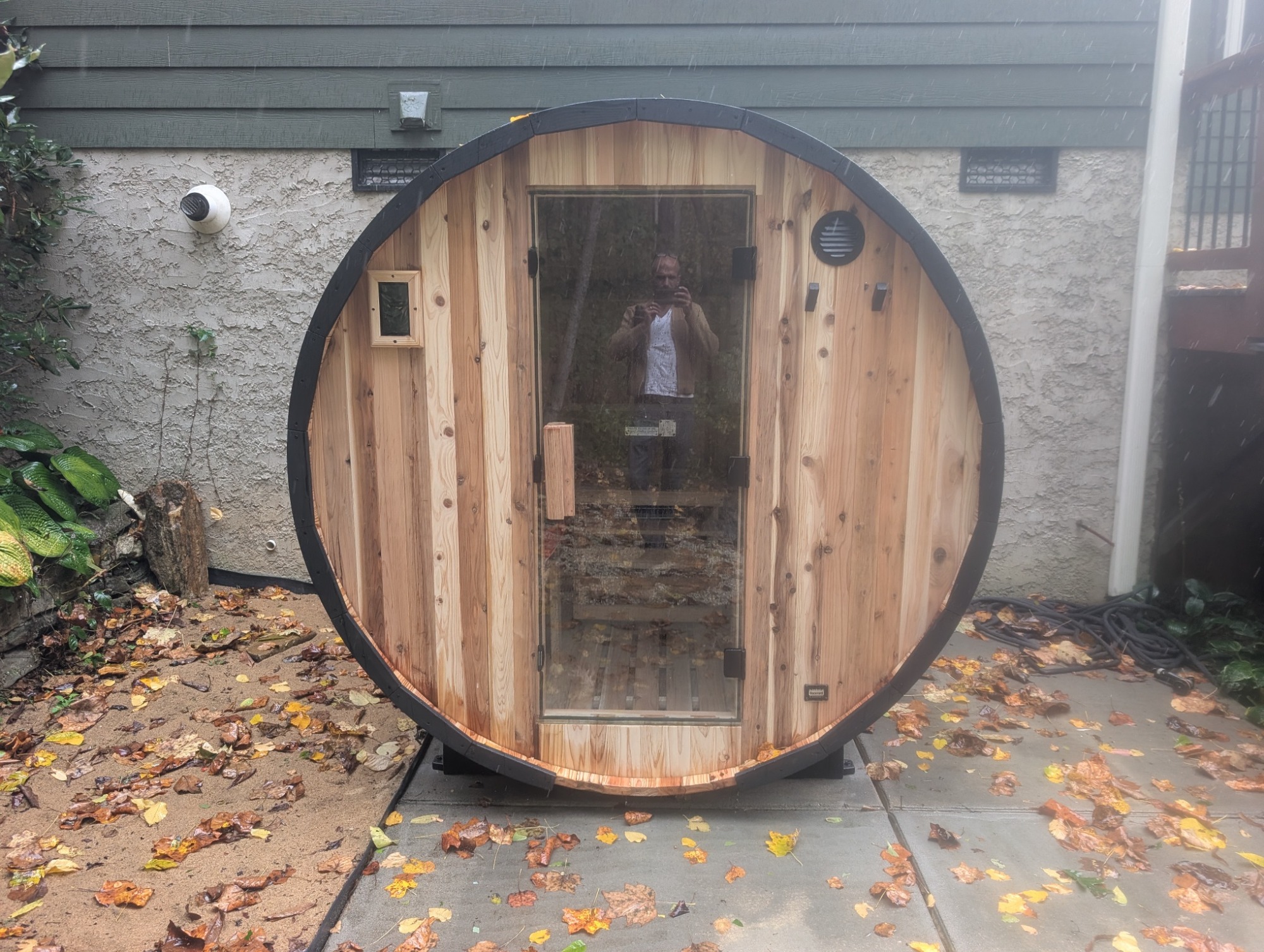 Finished wooden barrel sauna on a concrete pad against a house, rain in the air and autumn leaves on the ground. A person holding a phone is reflected in the glass door.